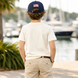 Person wearing a navy cap with an American flag and a white t-shirt, standing by a waterfront with boats in the background.