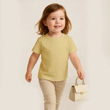 Child wearing a yellow shirt and beige pants holding a small cream-colored handbag against a plain background