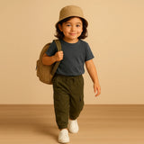 Child wearing a beige bucket hat, dark blue shirt, green pants, and white shoes, holding a brown backpack against a beige background.