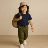 Child wearing a navy shirt, green pants, beige hat, and carrying a backpack on a beige background
